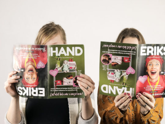 Två personer håller Erikshjälpens tidning framför sina ansikten. Tidningen går att läsa från två olika håll, och därför håller de tidningen på olika håll.