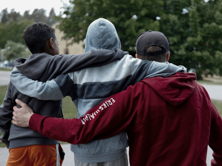 Ryggtavlor på tre killar som håller varandra över axlarna. Personen längst ut till höger har en Erikshjälpen-hoodie på sig.