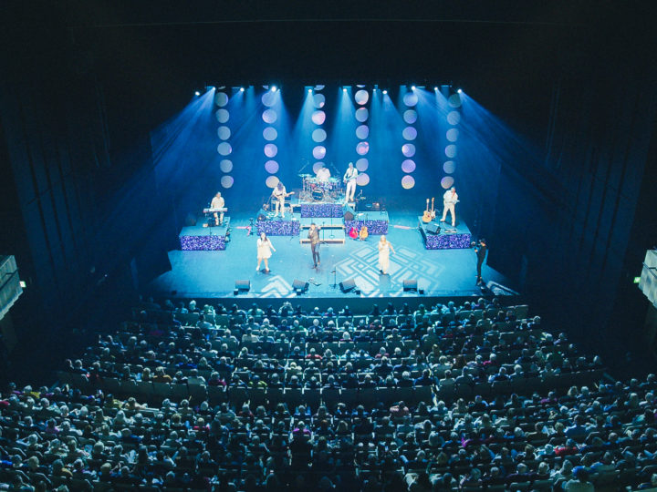 En artist med band står på scen i blått ljus. I publiken sitter mängder med människor.